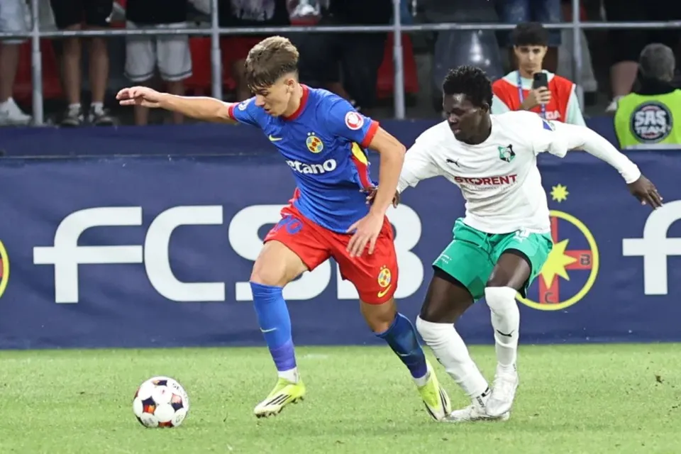 Un jucător de fotbal în echipament albastru și roșu dispută un balon cu un alt jucător îmbrăcat în alb și verde pe un teren de fotbal.