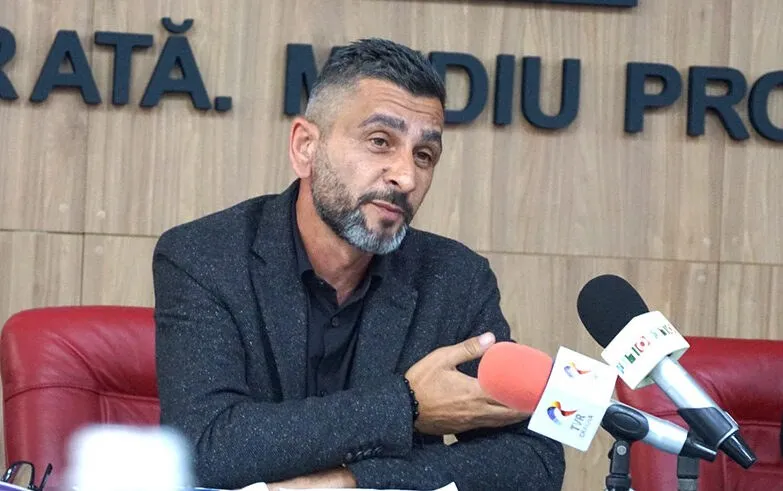 Un bărbat cu barbă și păr scurt stă la o masă în fața unor microfoane, purtând un sacou închis la culoare.