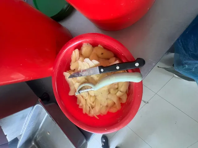 Un bol roșu conține bucăți de alimente tăiate, având un cuțit și un unghiator așezate deasupra.