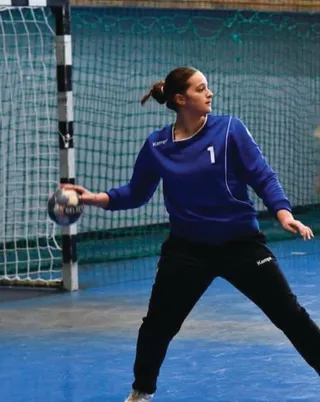 O femeie cu părul prins într-o coadă de cal poartă o cămașă albastră cu numărul unu și pantaloni negri, ținând o minge de handbal.