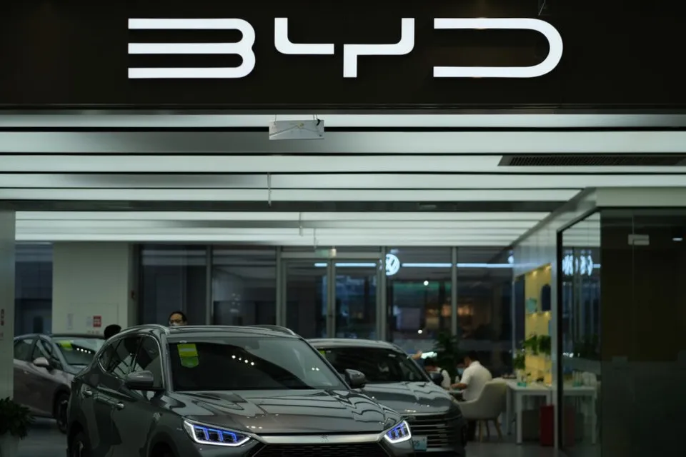 Două mașini de culoare gri sunt parcate în interiorul unui showroom sub un logo luminos alb pe un fundal negru.