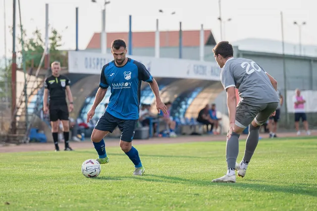 Un jucător de fotbal în echipament albastru conduce mingea pe un teren de iarbă verde în fața unui alt jucător în echipament gri.