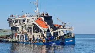 O navă mare de culoare albastră și albă cu o barcă de salvare portocalie suspendată pe lateral plutește pe apă sub un cer senin.