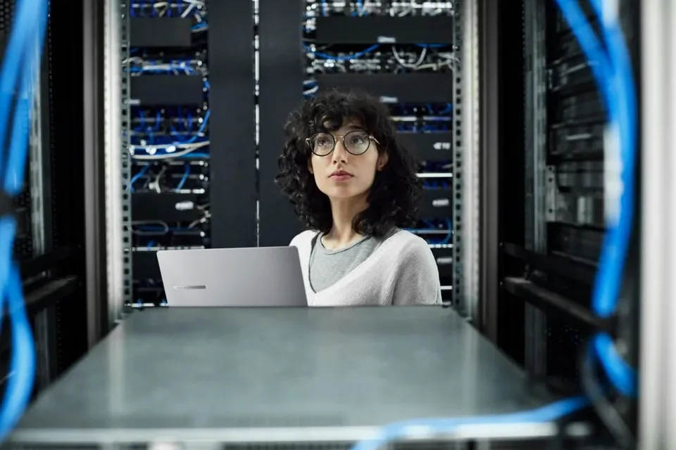 O femeie cu ochelari și părul creț stă la un birou cu un laptop în fața unor rack-uri de servere cu cabluri albastre.