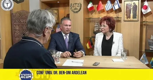 Trei persoane stau la o masă de lemn într-un studio, în fața unor steaguri și a unui panou cu diverse embleme pe perete.