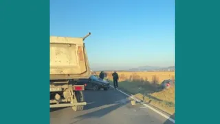 Un camion gri este parcat pe marginea unui drum asfaltat lângă un autoturism negru și un om care merge pe lângă vehicule.