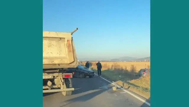 Un camion gri este parcat pe marginea unui drum asfaltat lângă un autoturism negru și un om care merge pe lângă vehicule.