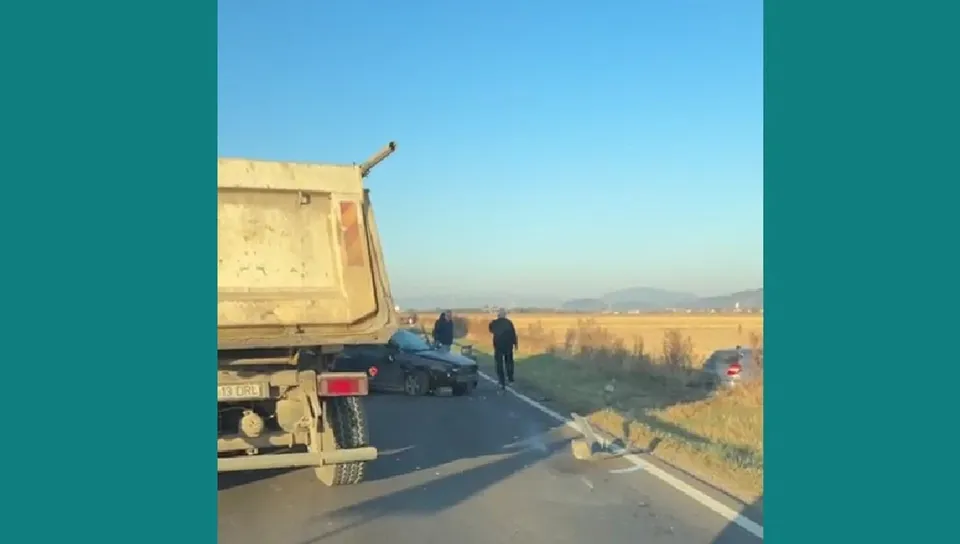 Un camion gri este parcat pe marginea unui drum asfaltat lângă un autoturism negru și un om care merge pe lângă vehicule.