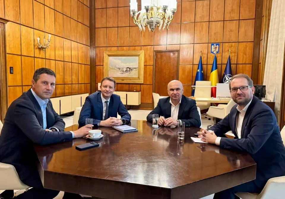 Patru bărbați în costume purtând cămăși deschise la guler stau așezați la o masă de lemn într-o încăpere cu pereți din panouri de lemn.