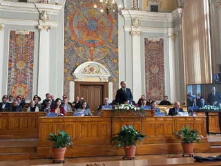 Oamenii stau așezați la mese lungi de lemn într-o sală înaltă cu panouri decorative și picturi murale mari pe pereți.