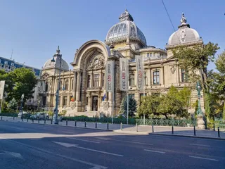 O clădire monumentală cu cupole și coloane este situată în spatele unor steaguri albe și a unor copaci, lângă un drum asfaltat cu marcaje albe.