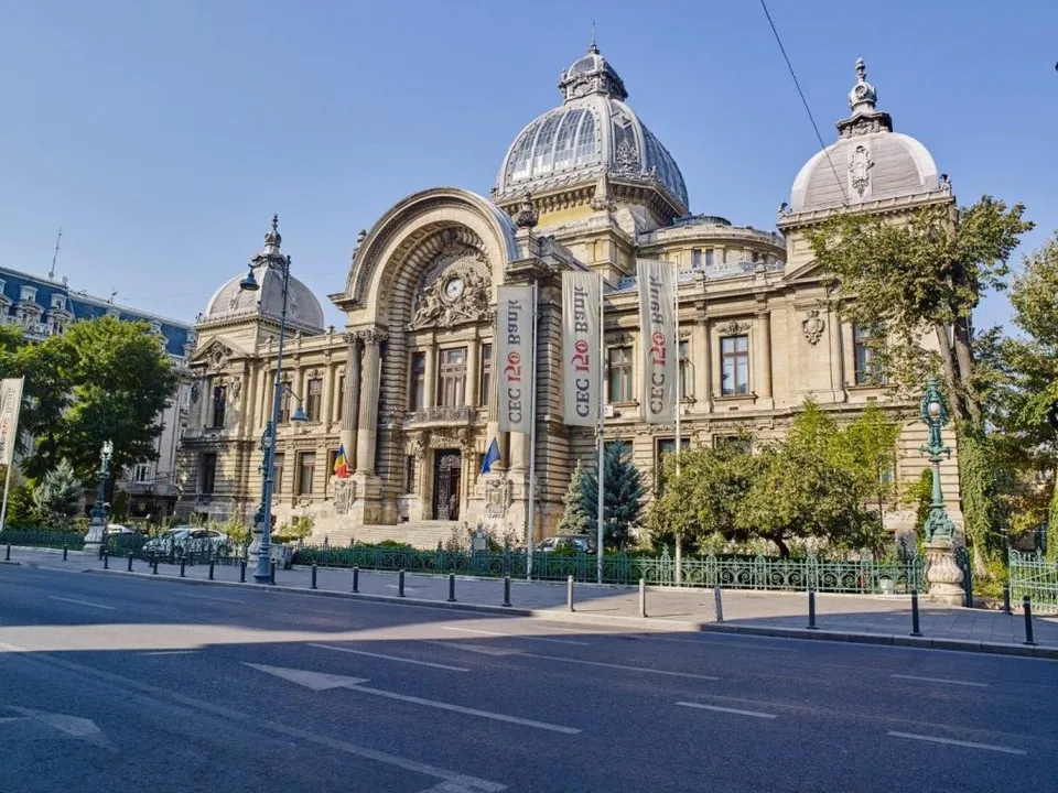 O clădire monumentală cu cupole și coloane este situată în spatele unor steaguri albe și a unor copaci, lângă un drum asfaltat cu marcaje albe.