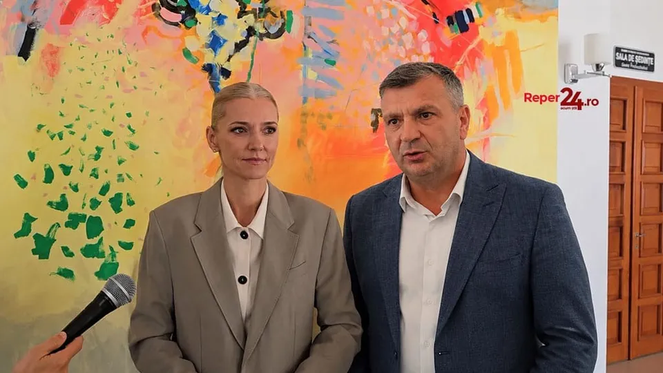 Un bărbat într-un sacou albastru și o femeie într-o jachetă gri stau în fața unui perete colorat lângă un microfon.