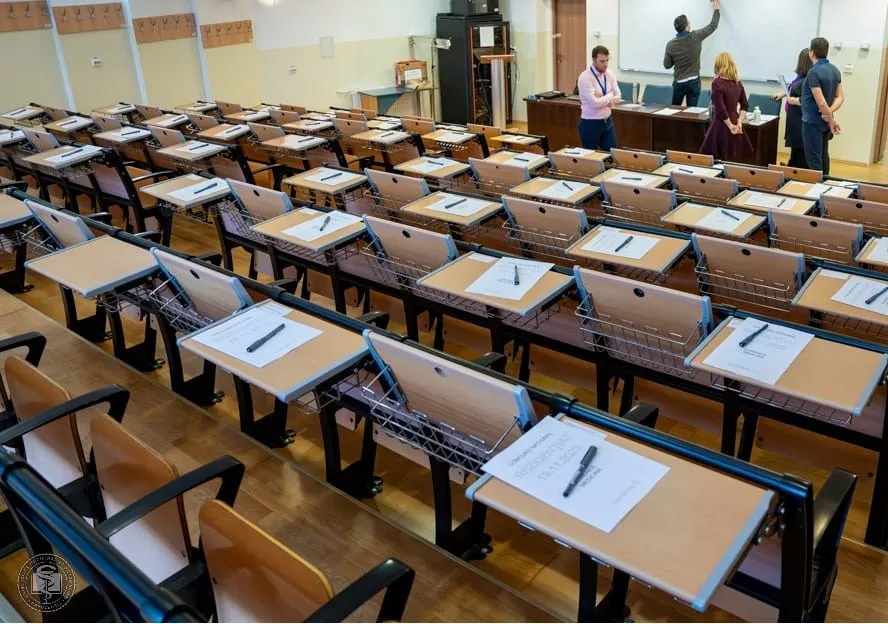 O sală de curs cu rânduri de birouri individuale dotate cu foi de hârtie și pixuri, în fundal fiind vizibile câteva persoane în picioare.