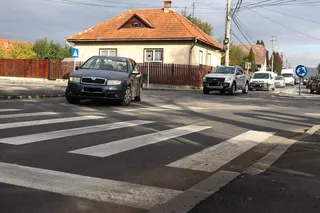 Un autoturism negru și un pick-up gri circulă pe un drum asfaltat lângă un punct de trecere a pietonilor marcate cu linii albe.