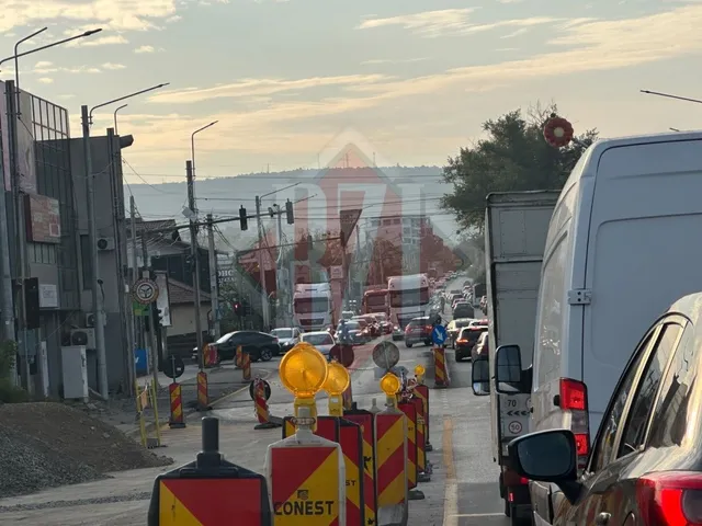 O serie de vehicule și utilaje de construcții sunt situate pe un drum lângă convoi de marcaje rutiere galbene și roșii.