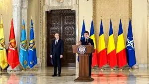 Un bărbat stă în spatele unui podium în fața mai multor steaguri colorate, în timp ce un alt bărbat stă în picioare în fundal.