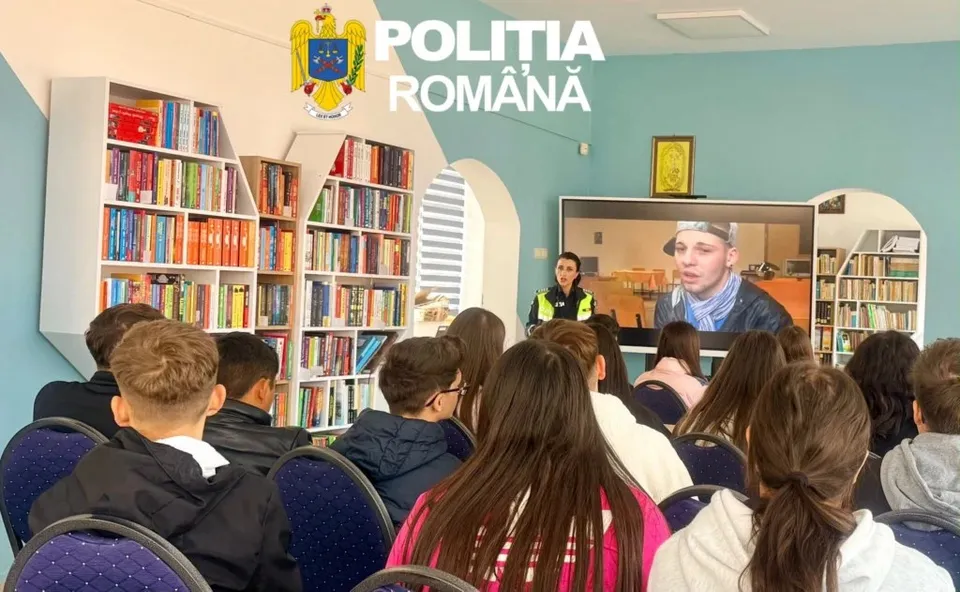 O grupă de copii stă pe scaune în fața unui ecran de televiziune care afișează două persoane într-o încăpere cu rafturi pline de cărți.