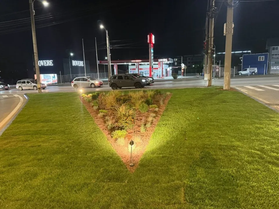 O insulă triunghiulară cu gazon, plante și corpuri de iluminat este mărginită de șosea, mașini, stâlpi și clădiri comerciale nocturne.