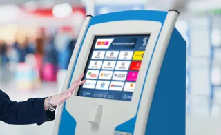 O mână de femeie atinge ecranul tactil al unui terminal bancar într-un spațiu interior iluminat.