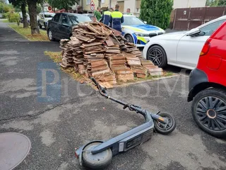 O trotinetă electrică neagră este întinsă pe asfaltul de lângă un morman de scânduri de lemn și câteva autoturisme parcate.