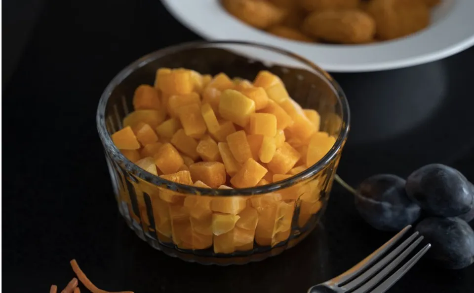Un bol de sticlă transparent conține cuburi de fructe sau legume de culoare portocalie, așezat pe o suprafață închisă lângă câteva fructe întregi și o furculiță.