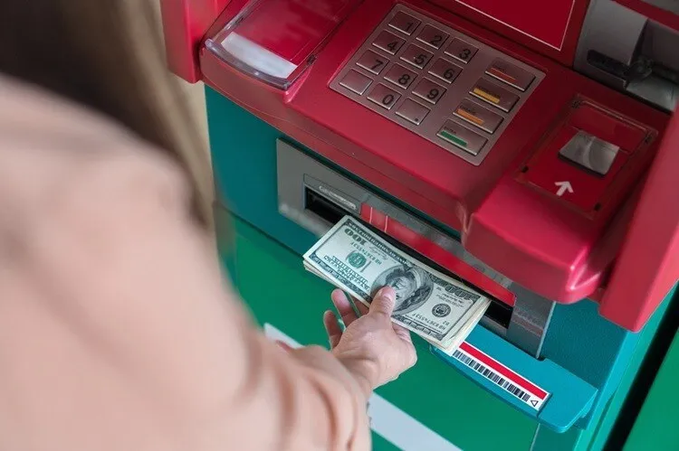 O persoană extrage bancnote de o sută de dolari dintr-un bancomat de culoare roșu și verde.