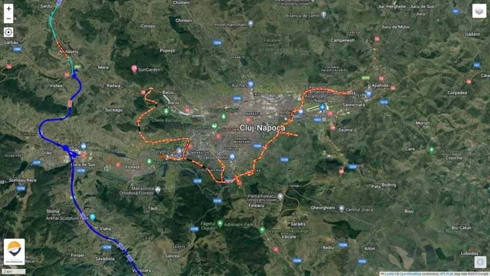 O hartă satelit de la înălțime arată o zonă urbană înconjurată de terenuri verzi, cu linii colorate marcate pe trasee rutiere.