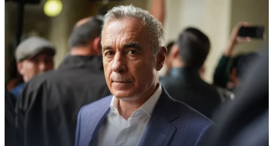 Un bărbat cu părul cărunt, purtând sacou albastru și cămașă albă, privește spre cameră în timp ce alte persoane stau în spatele lui.