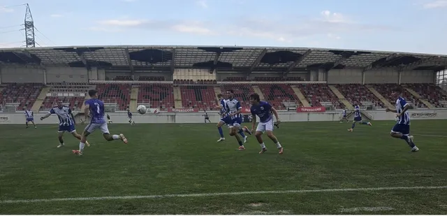 Câțiva jucători de fotbal în echipamente albastre și albe aleagă pe un teren de gazon verde sub un stadion cu tribune ocupate parțial.