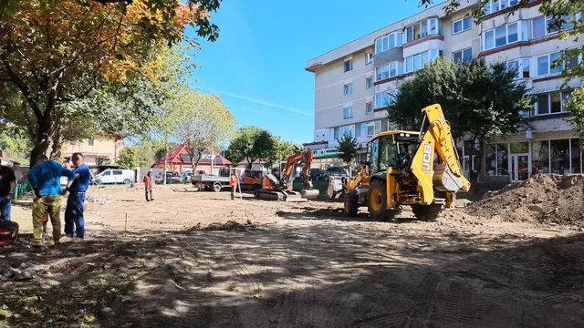 O excavatoare galbenă lucrează pe un teren de pământ lângă un bloc de apartamente, în timp ce câțiva oameni stau în apropiere.