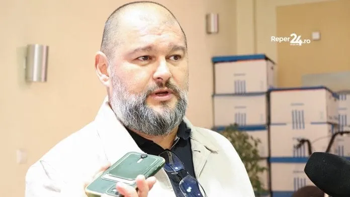 Un bărbat cu barbă și păr scurt, purtând o cămașă neagră și o sacoșă deschisă de culoare deschisă, ține un telefon mobil verde.