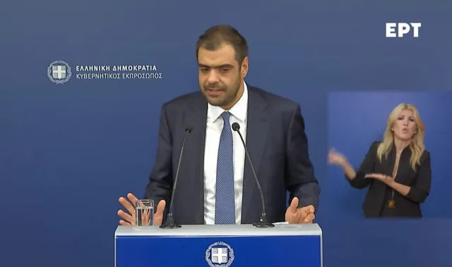 Un bărbat cu cămașă albă și cravată albastră stă în spatele unui podium alb, în timp ce o femeie cu părul blond este vizibilă într-un ecran secundar.