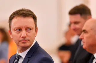 Un bărbat cu părul scurt și costum închis se uită spre lateral într-un spațiu cu fundal deschis și persoane în plan secund.