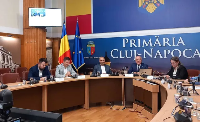 Cinci persoane stau la o masă curbată în fața unui panou albastru pe care scrie Primăria Cluj-Napoca, lângă steagurile României și Uniunii Europene.