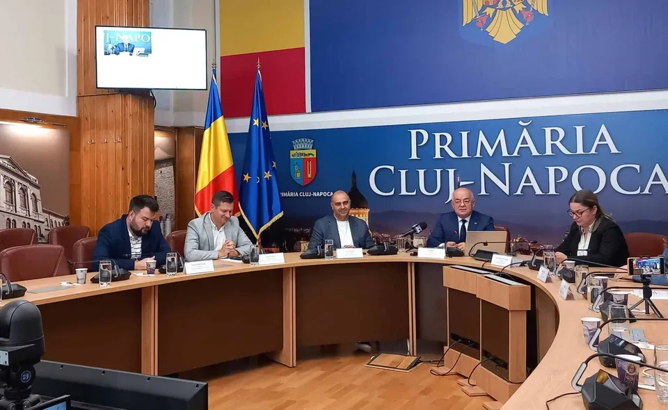 Cinci persoane stau la o masă curbată în fața unui panou albastru pe care scrie Primăria Cluj-Napoca, lângă steagurile României și Uniunii Europene.