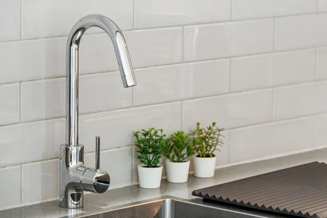 Un robinet metalic strălucitor este montat pe un blat de bucătărie lângă trei mici plante verzi în ghivece albe.