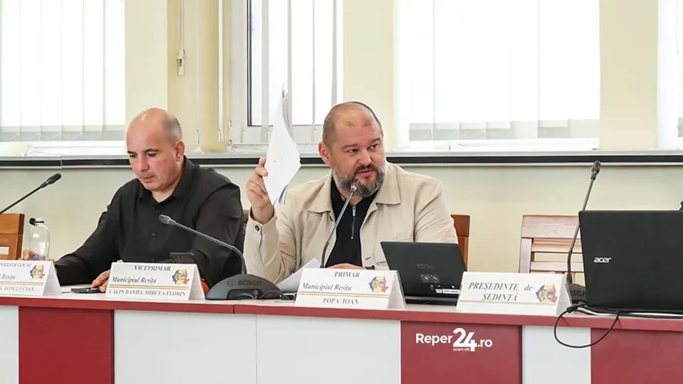 Un bărbat cu barbă și o jachetă deschisă ține o foaie de hârtie albă, stând la o masă lângă un laptop și un microfon.