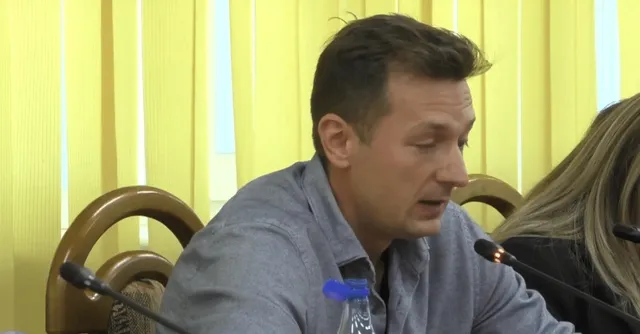 Un bărbat cu cămașă gri stă la o masă lângă un microfon, în fața unor jaluzele galbene.