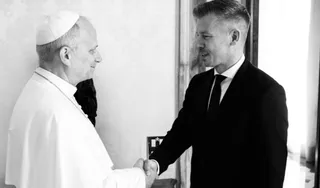 Un bărbat cu căciulă albă și haine albe strânge mâna unui bărbat într-un costum întunecat și cravată într-o încăpere luminoasă.