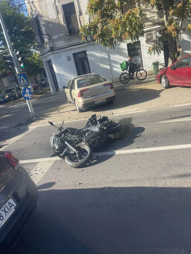 O motocicletă neagră este culcată pe asfaltul unei străzi lângă un autoturism de culoare aurie și un vehicul roșu.