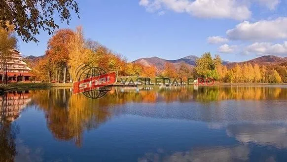 Un lac reflectă copacii cu frunze galbene și portocalii, un clădire maro și munții sub un cer albastru cu nori albi.