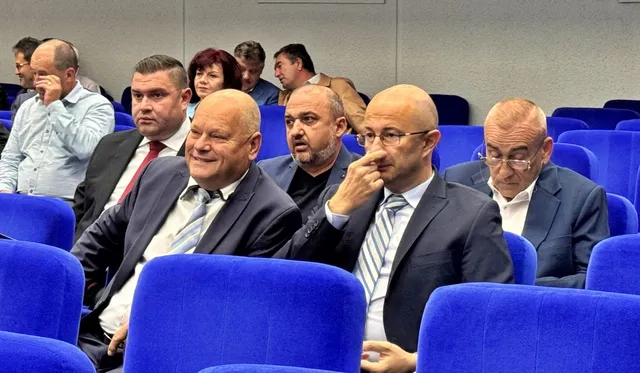 Un grup de bărbați în costume și cămăși stau așezați în scaune de culoare albastră într-o sală de conferințe.