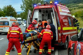Patru persoane în uniforme roșii și galbenii transportă o persoană pe o targă lângă un vehicul de intervenție de culoare roșie.