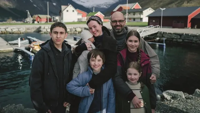Un grup de șapte persoane, inclusiv copii și adulți, pozează în fața unui lac cu case colorate și munți în fundal.