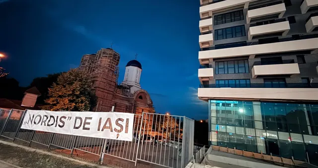 Un banner alb cu textul scris negru este atașat de un gard metalic, în fața unui bloc modern și a unei clădiri vechi sub cerul nopții.