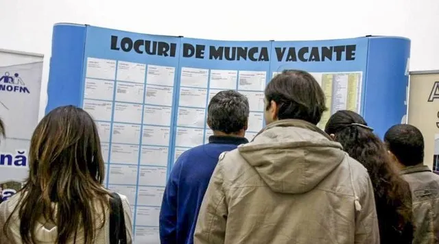 Oameni în haine groase privesc din spate un panou albastru cu afișe, sub un banner cu textul „LOCURI DE MUNCĂ VACANTE”.