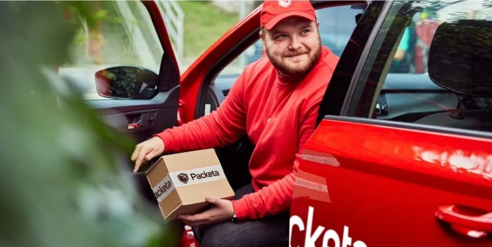 Un bărbat cu o căciulă roșie și un tricou roșu ține o cutie de carton cu inscripția Packeta lângă o mașină roșie.