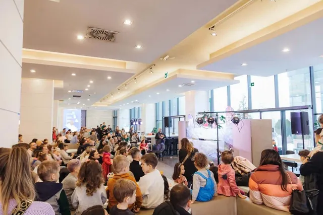 O mulțime de oameni stă așezată pe scaune într-un spațiu interior iluminat, orientată spre o scenă cu elemente de decor și echipamente audio.