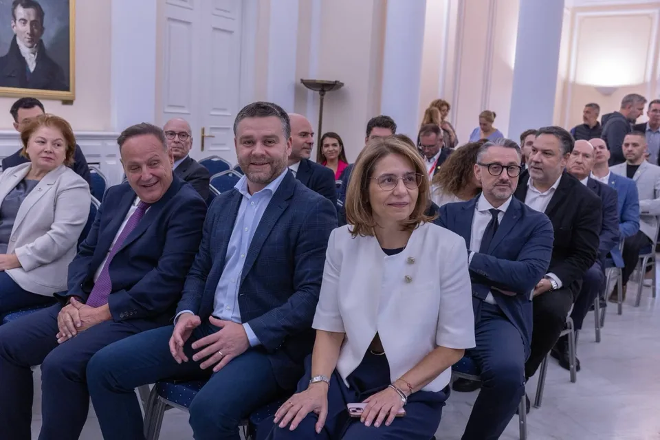 Un grup de persoane în haine elegante stă așezat pe scaune într-o încăpere cu pereți albi și un tablou încadrat pe perete.